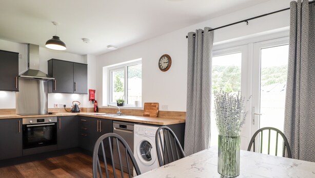 Cocina moderna con armarios oscuros, campana extractora de acero inoxidable, horno empotrado, lavavajillas y encimera de madera. Hay una mesa de comedor con mantel blanco y un jarrón de flores.