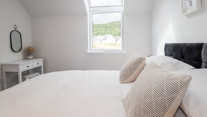 Un dormitorio con tragaluz, cama con ropa de cama blanca, mesita de noche con un florero y un cuadro en la pared.