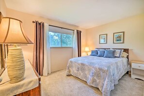 Un dormitorio con cama, mesita de noche, lámpara, ventana con cortinas y dos cuadros en la pared.