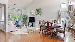 A spacious living room with a wooden dining table, comfortable seating, a flat-screen TV, and large windows offering natural light and views. - Wine Country Gateway w/ Views Btwn Napa & SF (Vallejo)