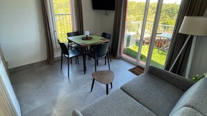 A modern living room with a grey sofa, a dining table with chairs, a TV mounted on the wall, and a balcony with a view of greenery.