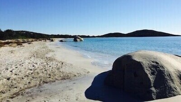 A beach with clear blue water, a large rock in the foreground, and a distant hill.