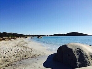 A beach with clear blue water, a large rock in the foreground, and a distant hill.