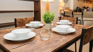 Una mesa de comedor dispuesta con platos, tazones y vasos blancos, junto a una pequeña planta en maceta y una lámpara.