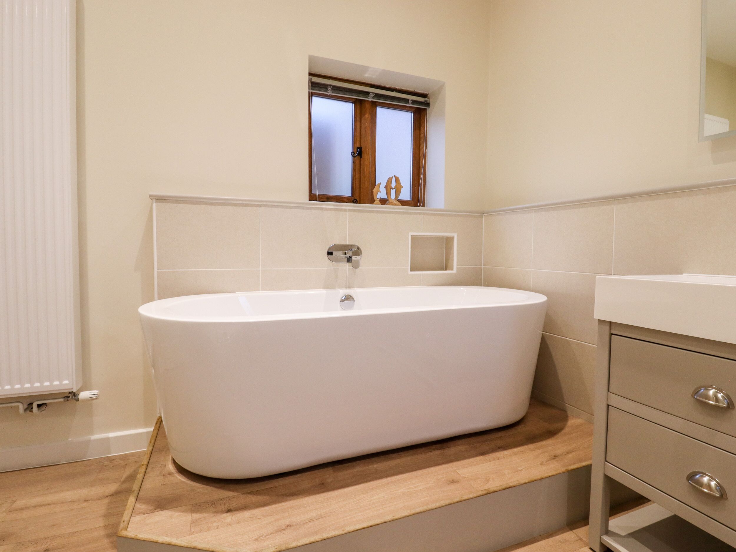 A modern bathroom with a freestanding bathtub, a wooden vanity with drawers, and a small window above the bathtub.