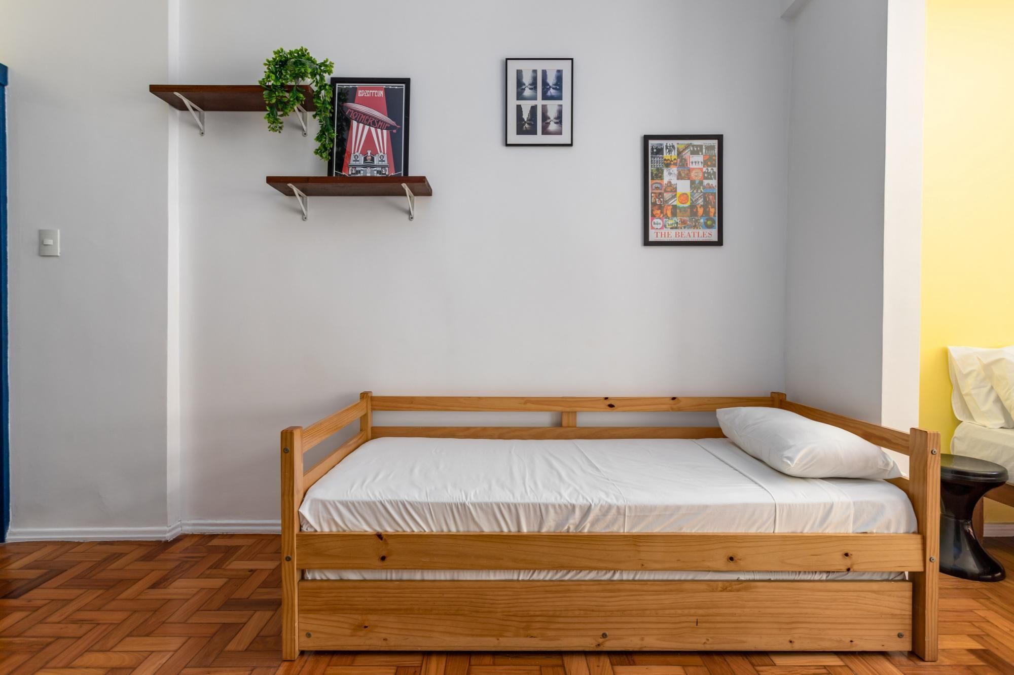 Uma cama de madeira simples com roupa de cama branca em um quarto com paredes brancas. Há duas imagens emolduradas na parede, uma de uma banda e outra de uma colagem. Uma pequena planta está em uma prateleira sobre a cama.