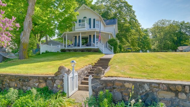 A two-story house with a front porch, a stone retaining wall, and a flag. - Elegant 1905 Home: Steps to Torch Lake & Dtwn (Alden)