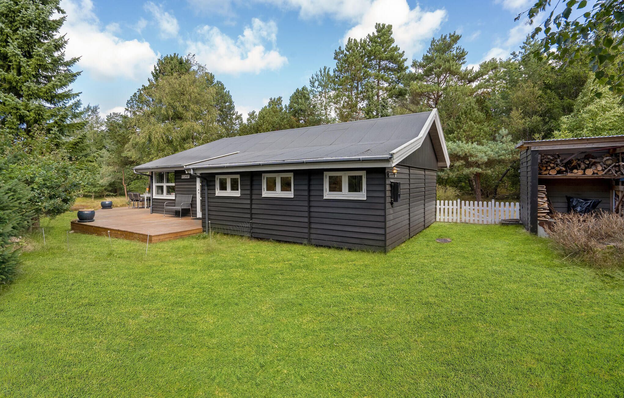 A small, dark-colored cabin with a deck, surrounded by a grassy yard and trees.