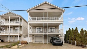 Un edificio residencial de dos plantas con un balcón en el piso superior, un garaje con un SUV negro estacionado adentro, y una fila de árboles de hoja perenne al fondo.