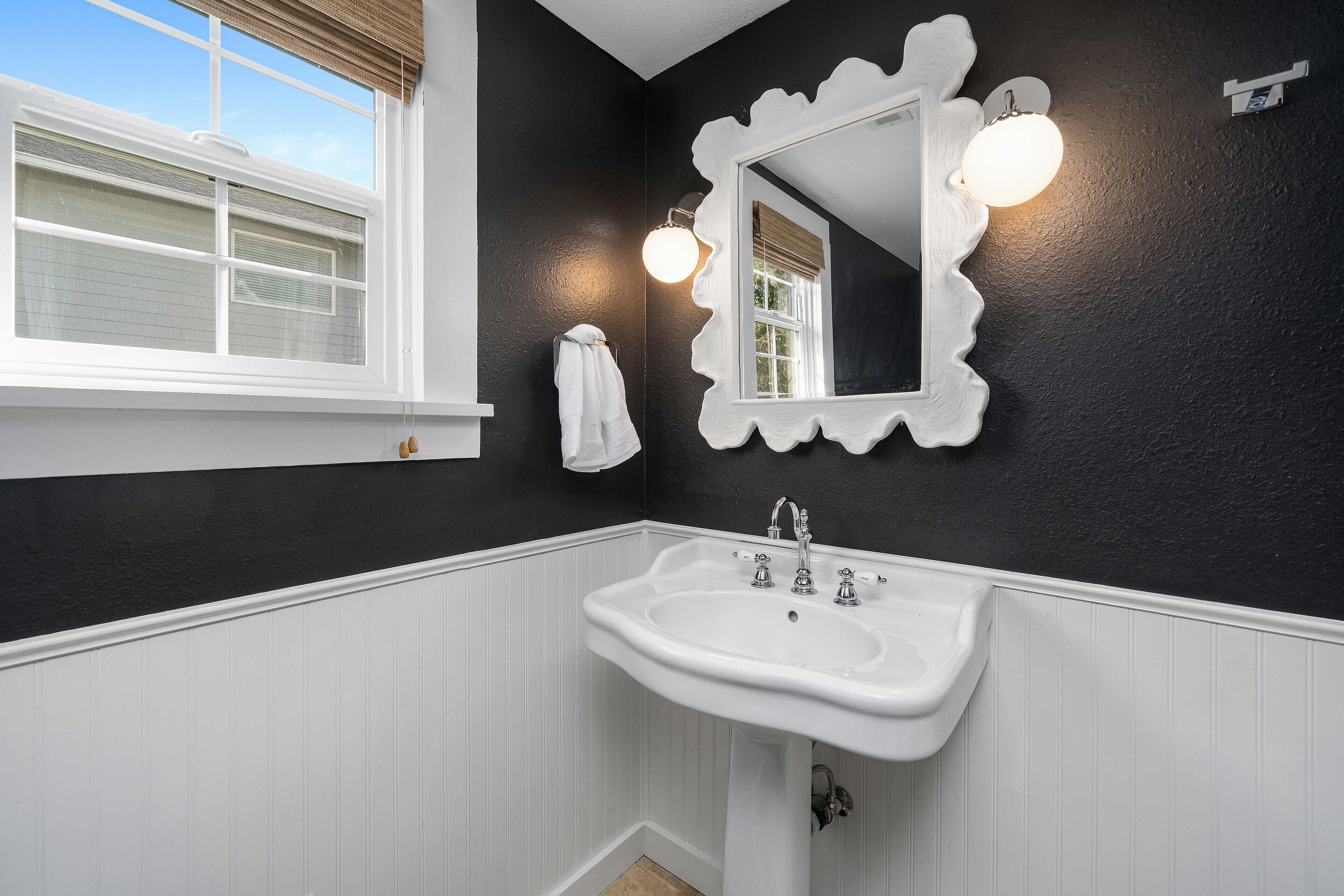 Une salle de bain avec un lavabo blanc, un miroir doté d’un cadre à motifs festonnés et une fenêtre avec des stores.