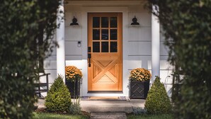 A wooden front door with a window above it, flanked by two potted plants with yellow flowers.