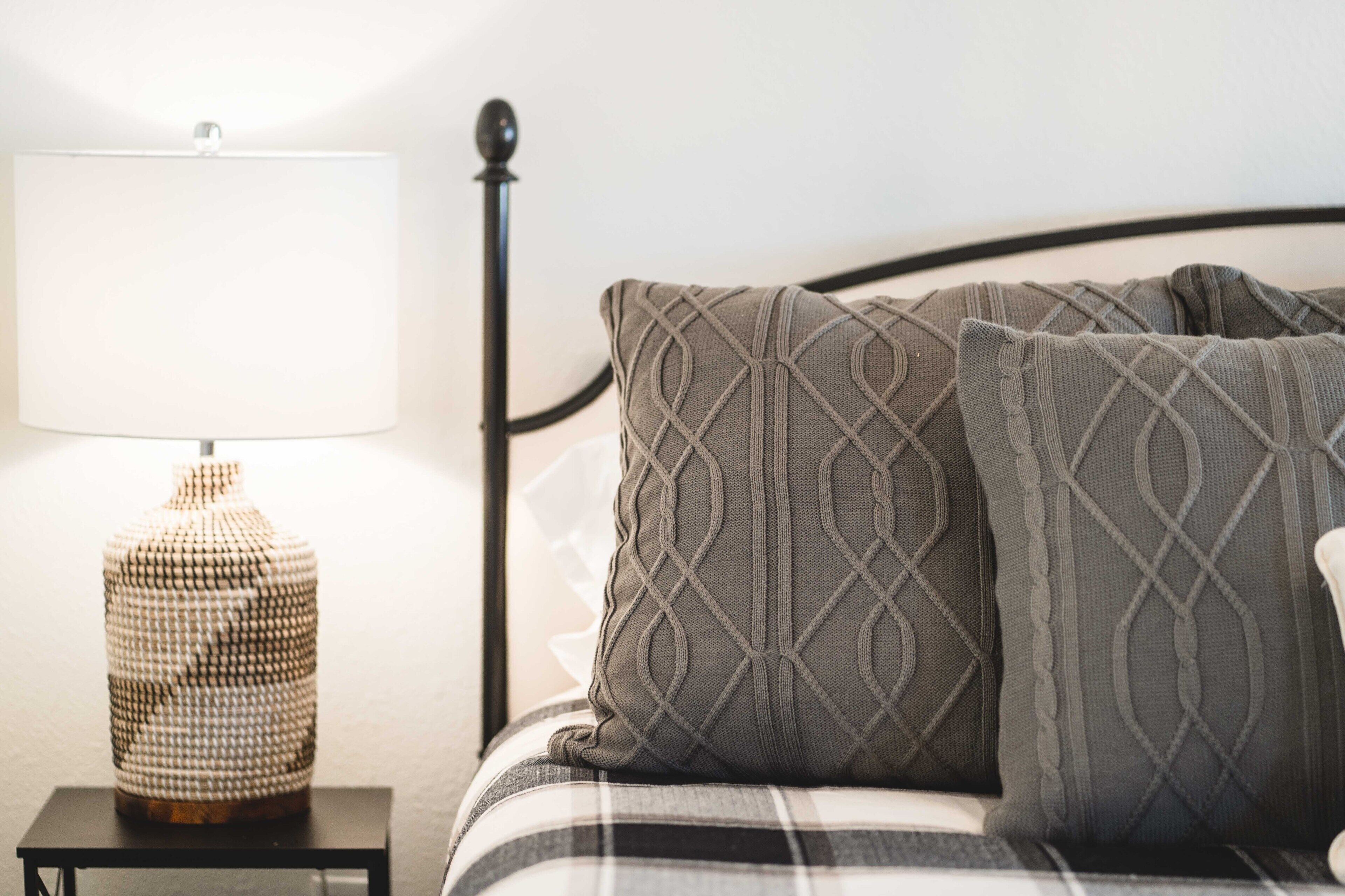A neatly made bed with a plaid blanket and patterned pillows, a bedside table with a lamp, and a decorative vase.