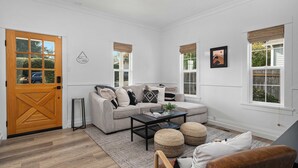 A living room with a sofa, coffee table, and ottomans, a wooden door, and windows with blinds.