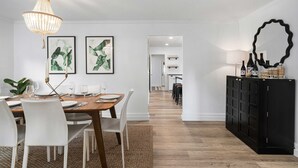 A dining area with a wooden table, white chairs, and a chandelier. There are framed artworks on the wall and a black cabinet with decorative items.