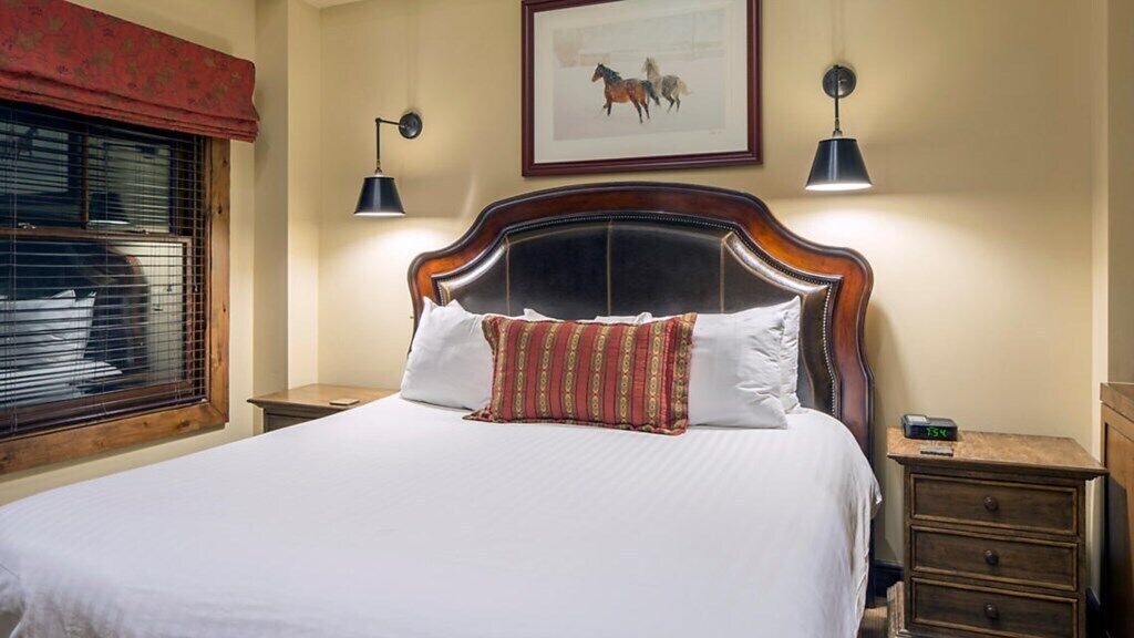 A bed with a wooden headboard, two bedside lamps, a clock, and a framed picture on the wall.