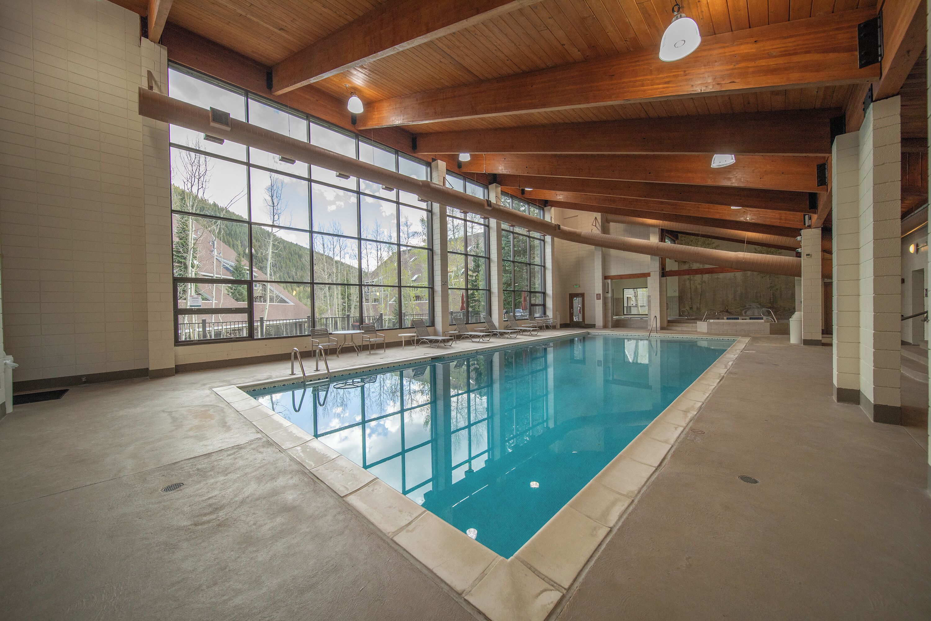 Une piscine intérieure avec vue sur les montagnes grâce à de grandes fenêtres, des poutres en bois au plafond et un sol carrelé.