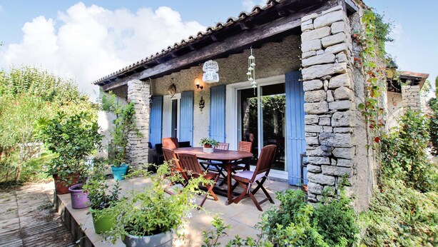 Un bâtiment en pierre avec un toit en tuiles, un patio couvert avec une table et des chaises, et des plantes en pot.