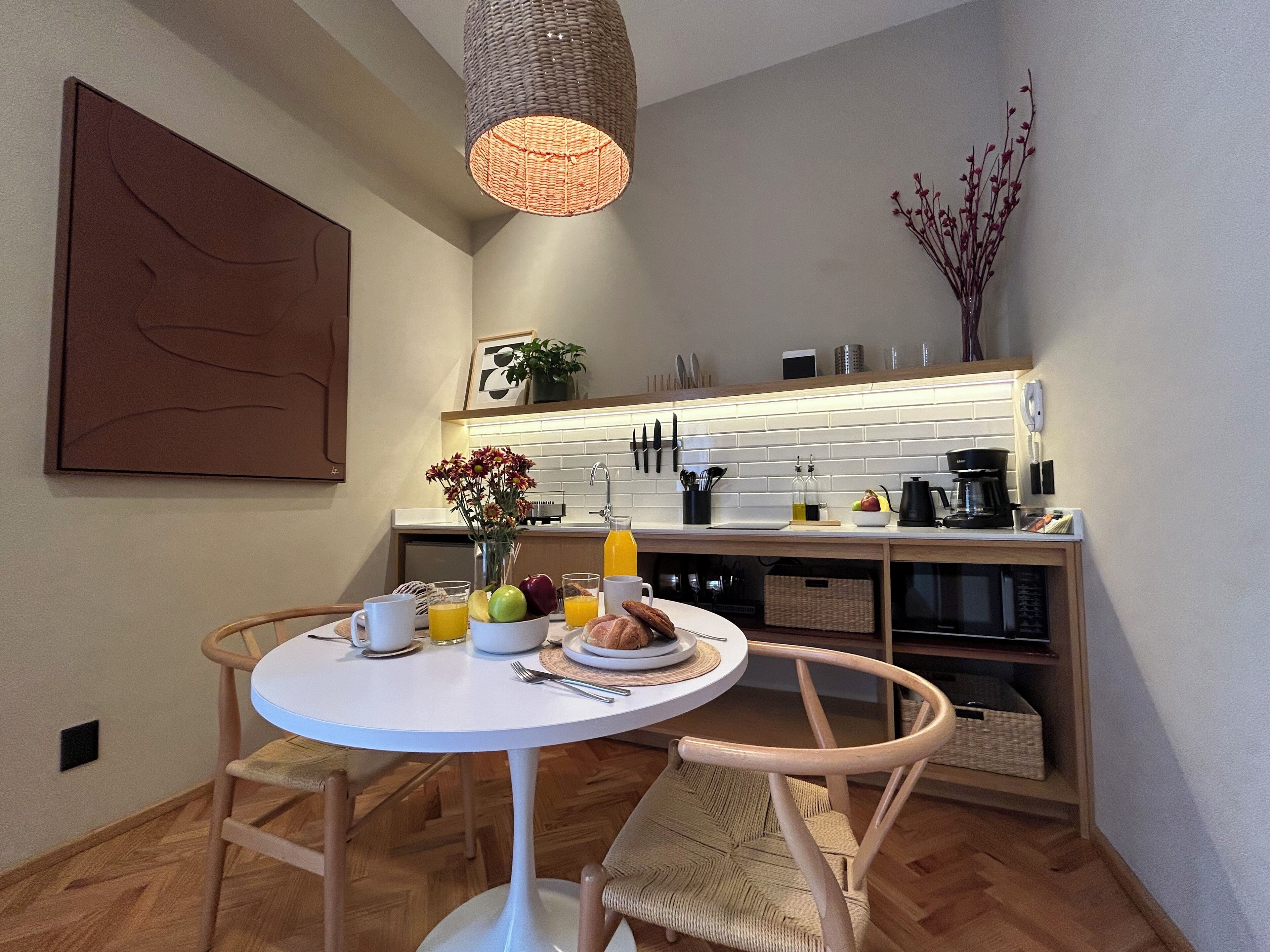 A compact kitchen with a dining area, featuring a round table, two chairs, a hanging light fixture, and a small potted plant.