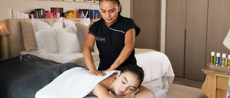 En massagebehandling i ett rum med en säng, kuddar, ett nattduksbord med flaskor och en vägg med inramade bilder.