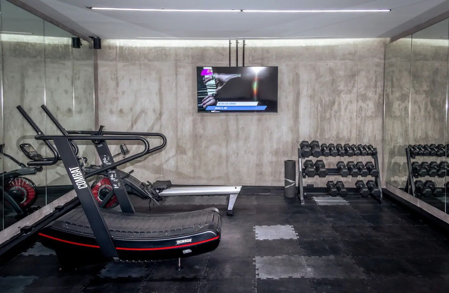 Een sportschool met cardioapparatuur, een televisie en een rek met gewichten.