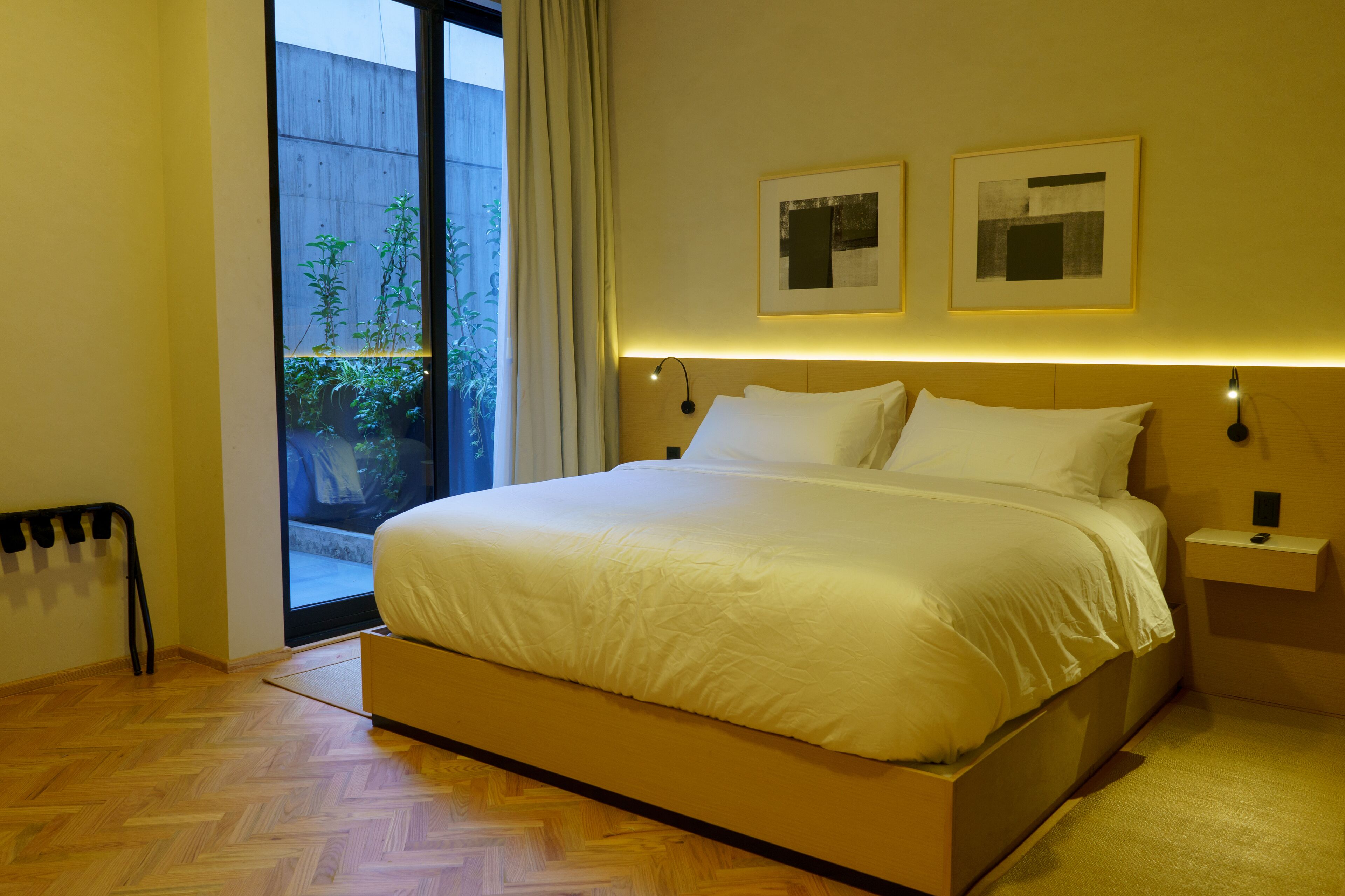 A bedroom with a large bed, a nightstand, two wall-mounted lamps, and a window with a view of greenery.