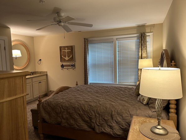 Une chambre à coucher comprenant un lit, un ventilateur de plafond, une fenêtre avec des stores, une table de chevet avec une lampe et une commode.