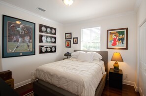 A bedroom with a bed, framed sports memorabilia, a nightstand with a lamp, and a window with blinds. - Beautiful Spanish Style Home In The Heart of Coral Gables (Coral Gables)