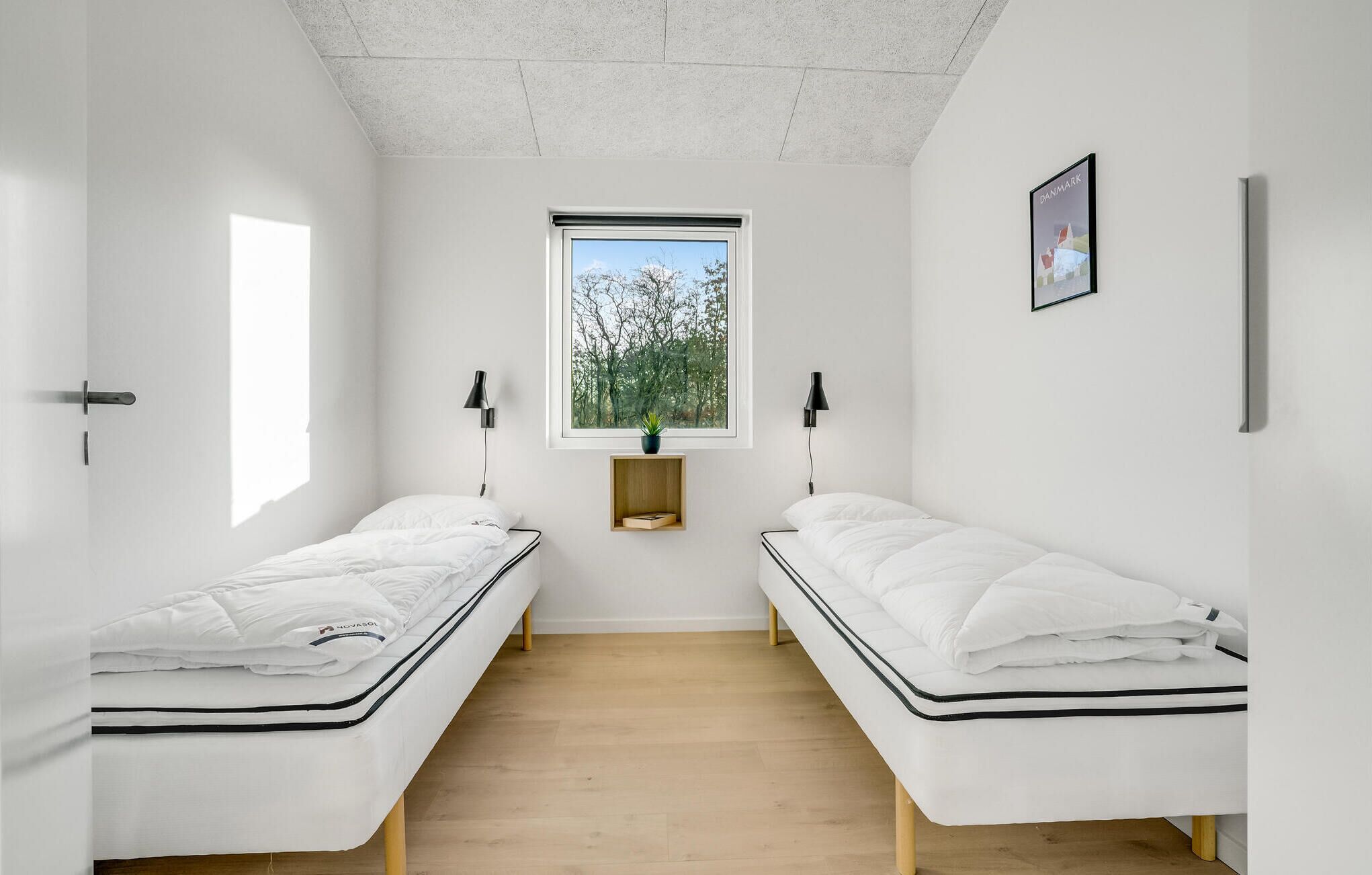 Een minimalistisch ingerichte kamer met twee eenpersoonsbedden, een raam met uitzicht op bomen, een klein plankje met een plant en een lijst aan de muur.