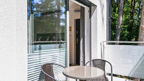 Een terras buiten met een ronde tafel en rieten stoelen, een glazen deur naar een binnenruimte en uitzicht op bomen en lucht.