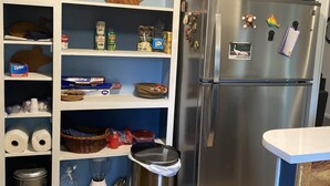 Ett kök med ett kylskåp i rostfritt stål, hyllor med olika livsmedel, en papperskorg och ett handfat.