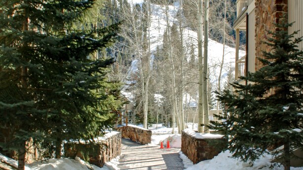 A paved pathway flanked by snow-covered ground and evergreen trees, leading towards a building with a brick facade. - Chateaux Dumont 2717 (Keystone)