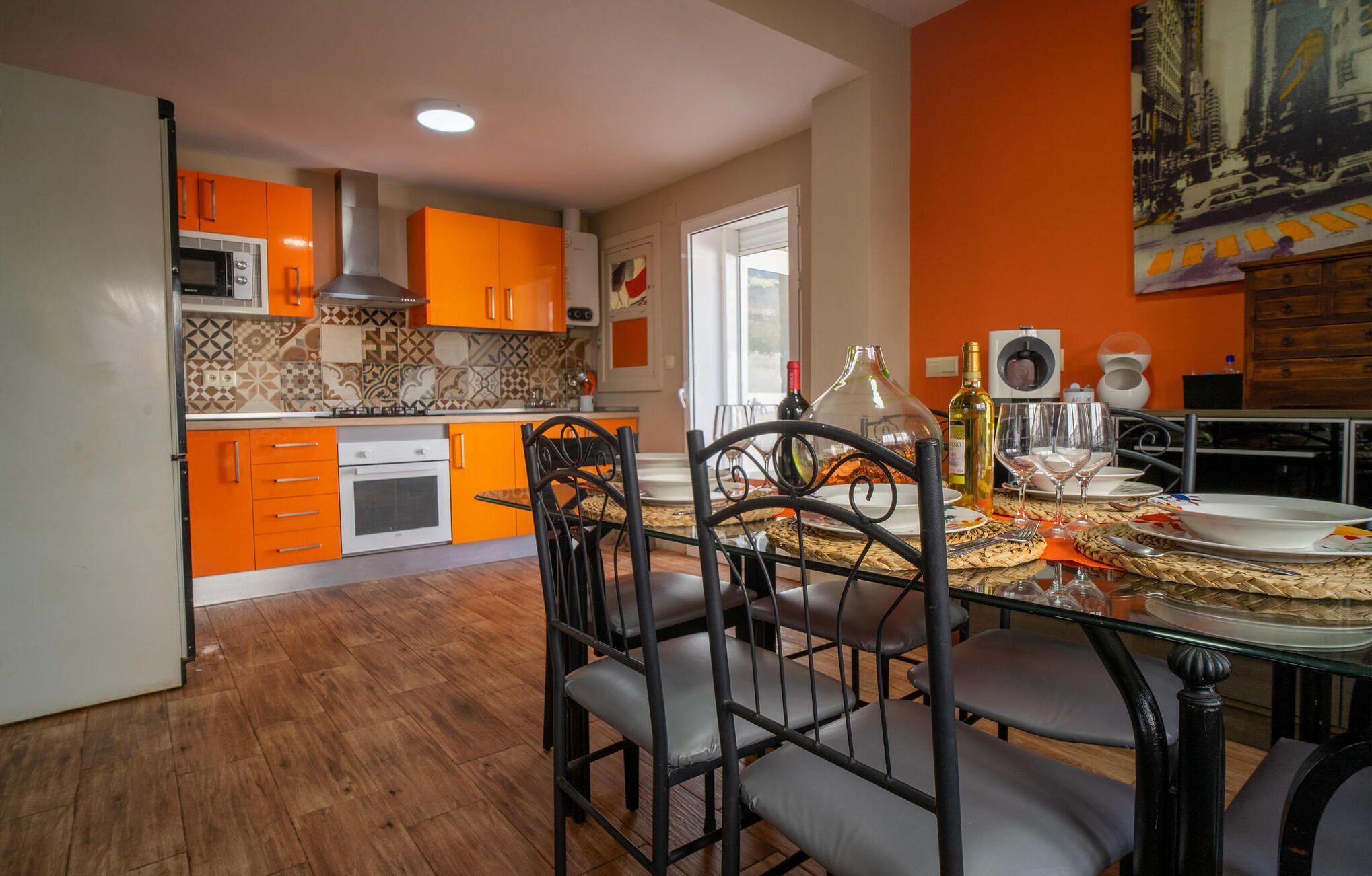 Uma cozinha moderna com armários cor de laranja, uma área de jantar com mesa de vidro e cadeiras pretas, e um mural com vista para a cidade na parede.
