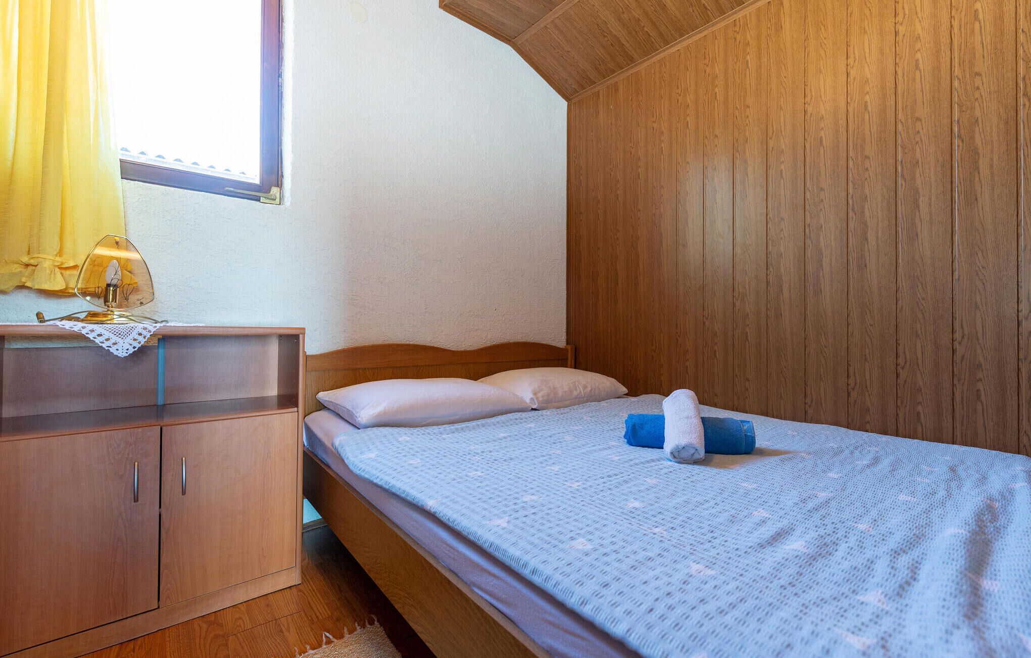 Une chambre avec une armoire en bois, un lit recouvert d’une couverture bleue et une fenêtre ornée de rideaux jaunes.