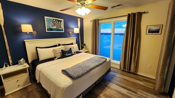 A bedroom with a large bed, two bedside tables, a ceiling fan, a window with curtains, and a view of the water. - Amazing Lake Views ~ 2 Kings ~ Outdoor Pool (Kimberling City)