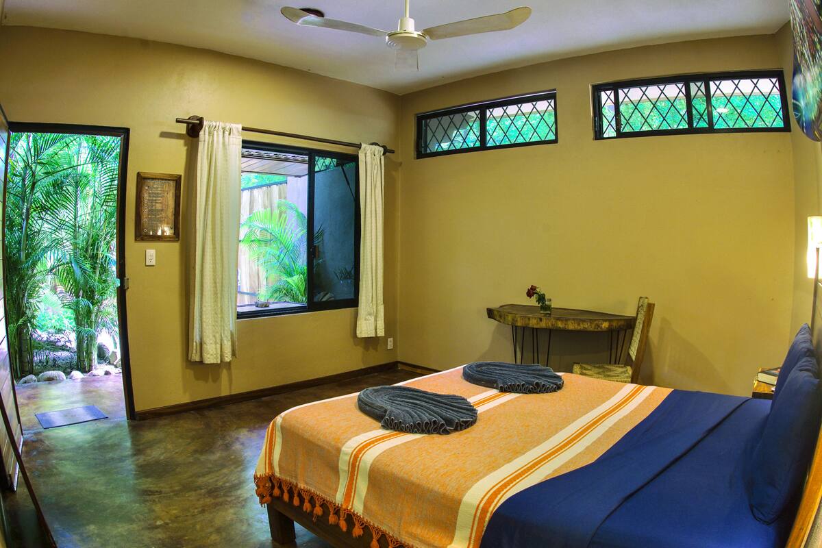 Quarto com cama, ventilador de teto, janela com cortinas, uma mesinha com um vaso e vista para o verde lá fora.