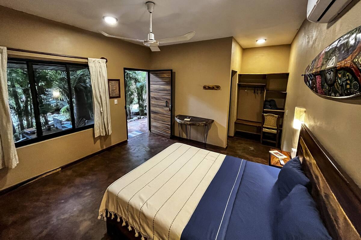 Quarto com cama, ventilador de teto, janela com cortinas e porta para uma varanda.