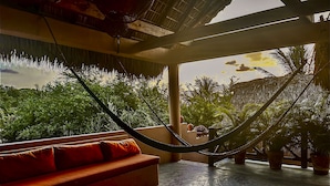 A hammock in a room with a view of lush greenery and a thatched roof structure. - Hotel Gallo de Mar (Puerto Escondido)