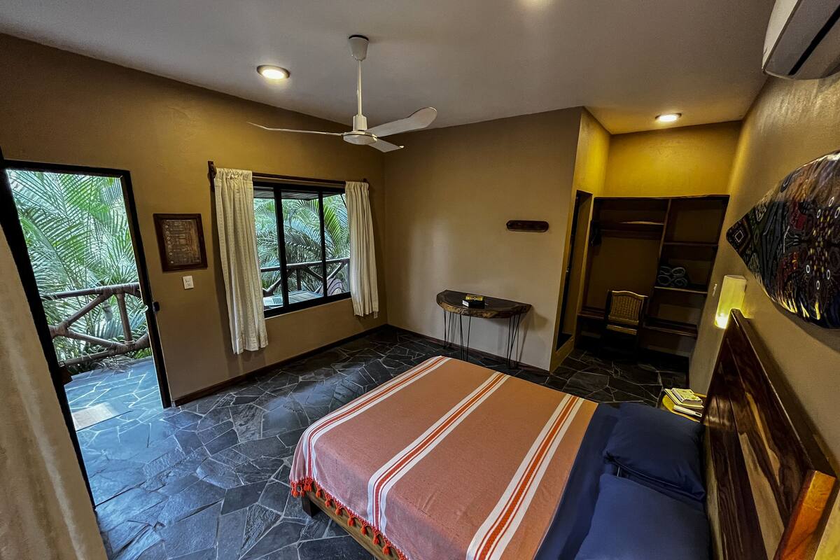 Quarto com cama, ventoinha no teto, janela com cortinas, uma mesinha e porta que dá para uma varanda.