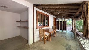 A room with a wooden dining table and chairs, a window with a wooden frame, and a thatched roof structure.