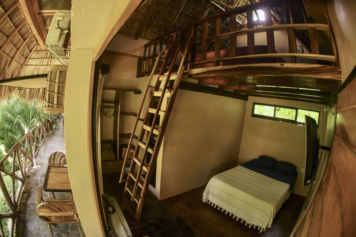 Quarto com cama, escada de madeira, janela e varanda com cadeiras de madeira.