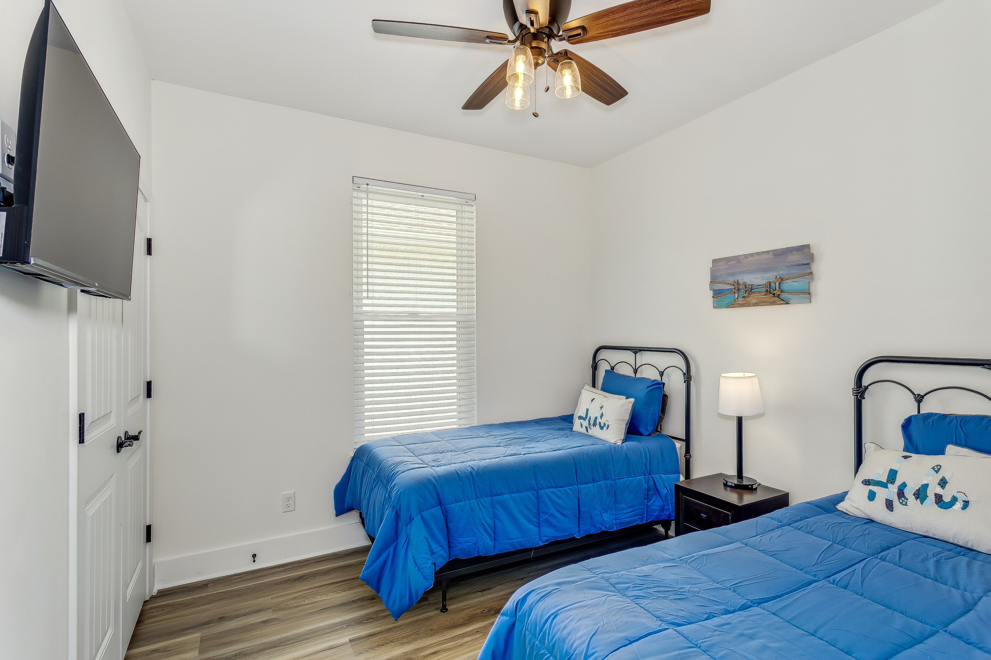A bedroom with two beds, a ceiling fan, a window with blinds, a painting, and a TV mounted on the wall.