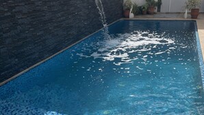En swimmingpool med et vandfald, omgivet af en flisbelagt terrasse og plantekasser.