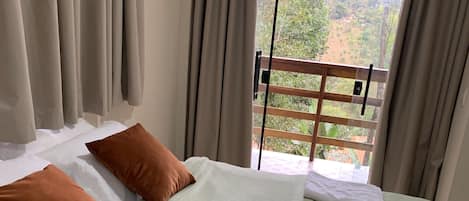 A hotel room with a bed, beige curtains, and a view of greenery and hills.