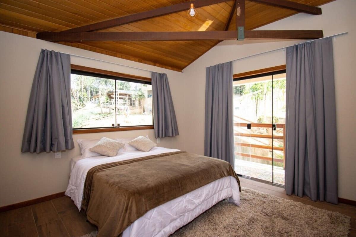 Quarto com cama grande, vigas de madeira no teto e vista através de uma porta de vidro deslizante.