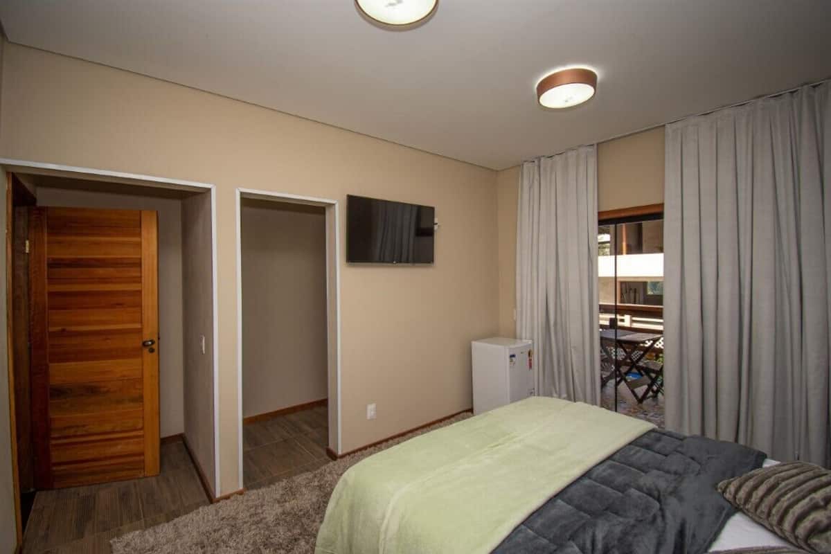 Quarto com cama, televisão, um pequeno frigorífico e varanda com cortinas.