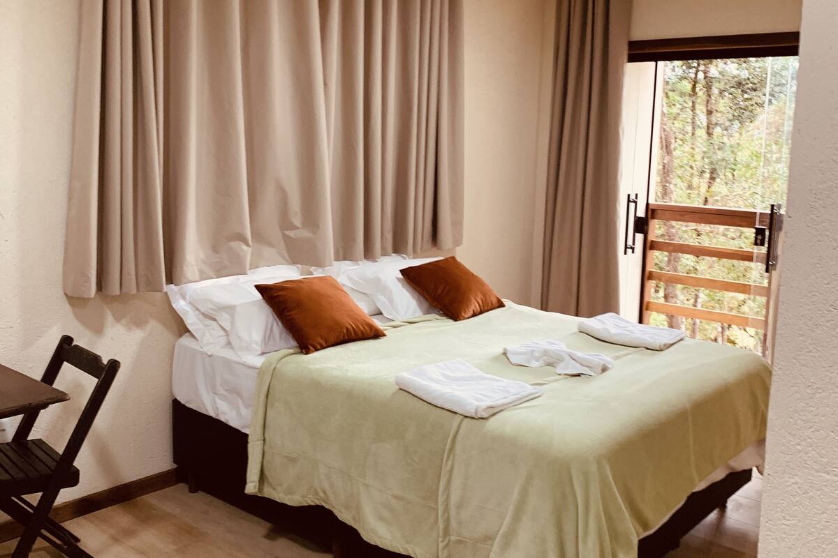 Uma cama bem arrumada com colcha verde e duas almofadas castanhas, uma cadeira de madeira e uma janela com cortinas.
