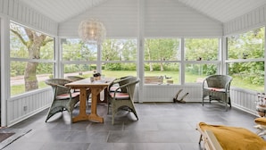 A covered patio with a wooden table and wicker chairs, large windows offering a view of a garden. - Nice home in Hemmet with WiFi (Hemmet)