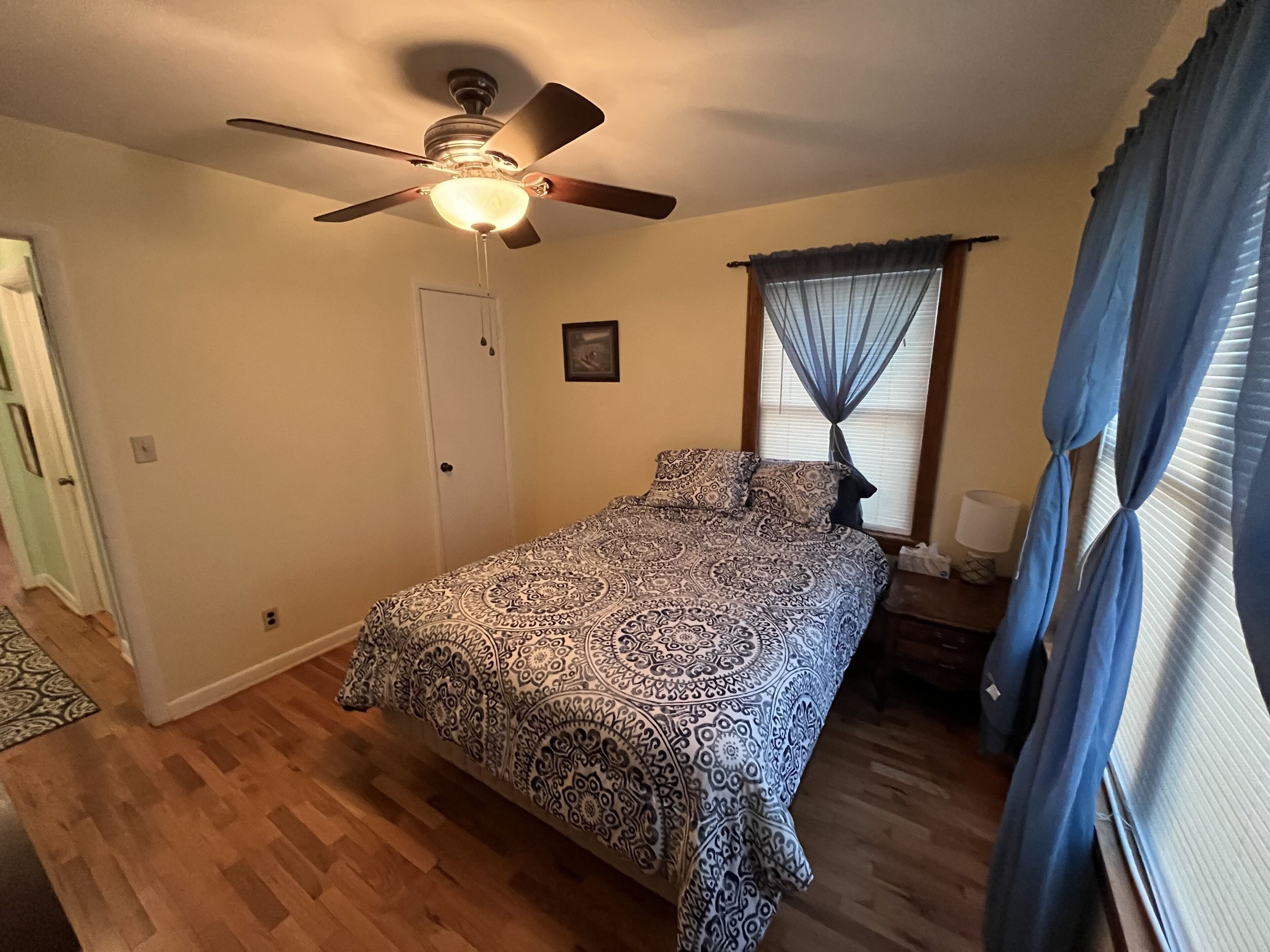 A bedroom with a bed, a ceiling fan, a window with curtains, and a door.