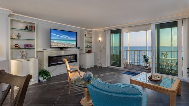 A modern living room with a flat-screen TV, a fireplace, and a balcony with a view of the sea. - Crystal Sands Beach Elegance (Siesta Key)