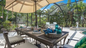 Uma área de refeições ao ar livre com uma mesa posta para dois, rodeada de cadeiras, sob um grande guarda-sol.